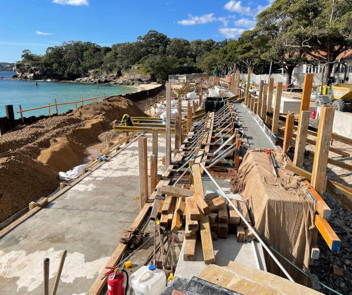 Construction with wooden posts, slabs of concrete and machinery visible against the backdrop of a calm turquoise bay with a thickly treed shoreline.