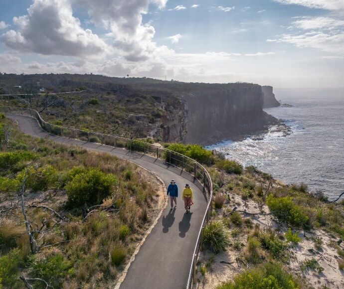 Aerial view of a couple on the Fairfax Walk at North Head, Sydney Harbour National Park, featuring a paved pathway with scenic ocean views and native vegetation.