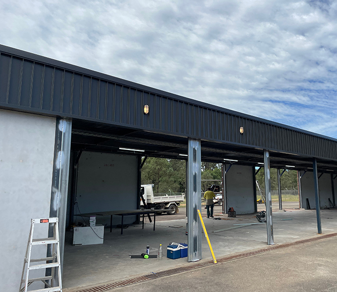 A partially constructed building with an open front, showing several workstations inside. Construction materials and tools are scattered around, and a worker in a yellow shirt is seen working inside. A white truck is parked in the background. The sky is cloudy.