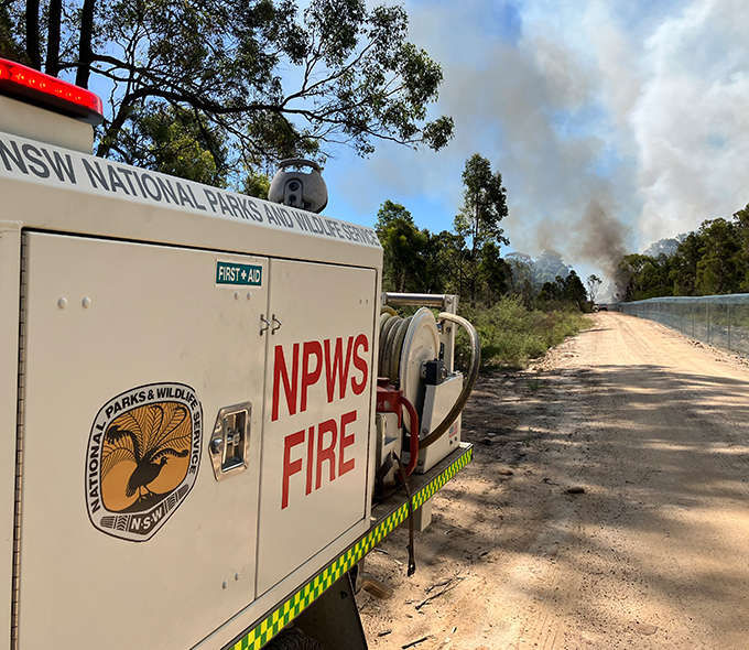 The image depicts a scene of a fire emergency in New South Wales, Australia. In the foreground, there’s a NSW National Parks and Wildlife Service (NPWS) fire truck parked on a dirt road. The side of the truck is labeled “NPWS FIRE” and also has a “FIRST-AID” logo. Equipment, including hoses for firefighting, is mounted on the side of the truck. In the background, smoke rises into the sky, indicating an ongoing wildfire. The natural environment features trees lining both sides of the dirt road.