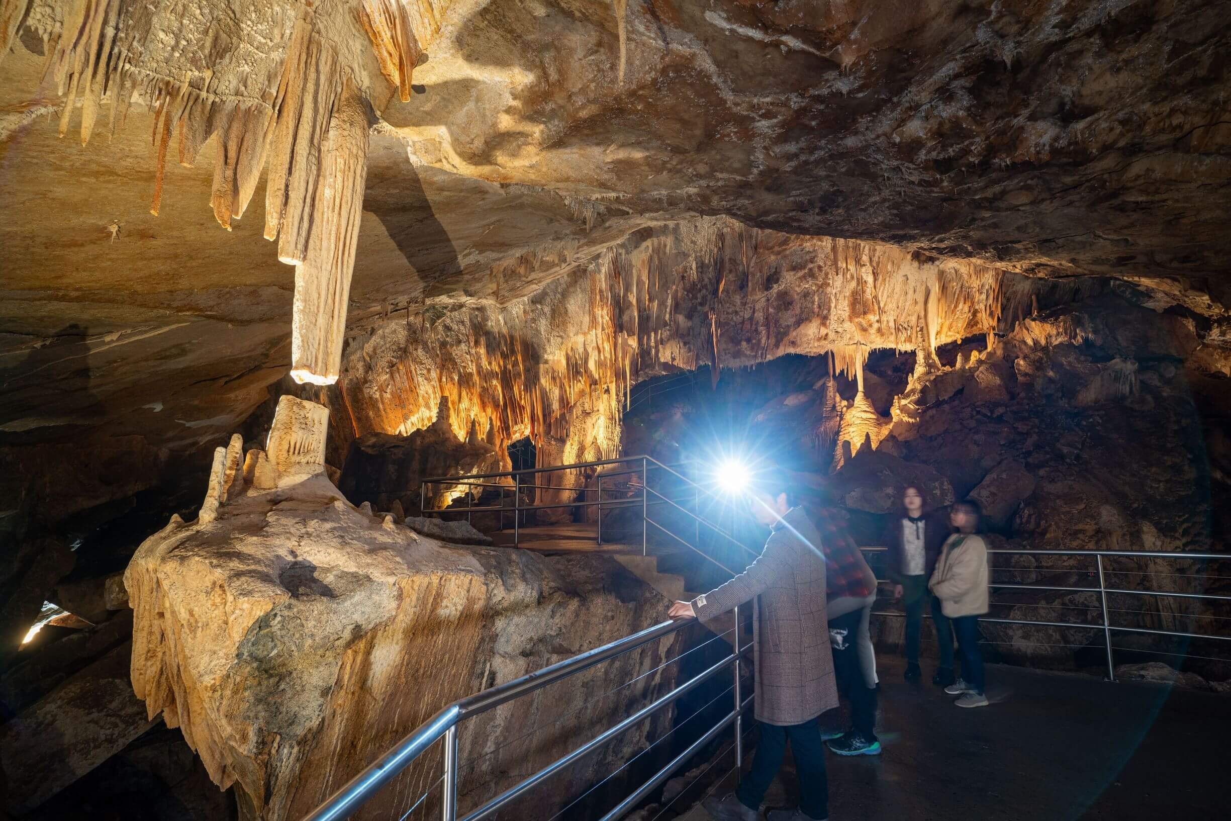 A group of people explore a cave with impressive stalactites and stalagmites. The cave is well-lit, showcasing intricate rock formations. A person in the foreground uses a flashlight to illuminate the cave walls. Metal railings guide the path through the cave.