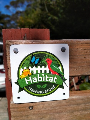 Sign attached to a wooden fence post featuring a circular logo with a white picket fence, a blue butterfly, a red bird, and a yellow frog. The text on the sign reads 'Habitat Stepping Stone.