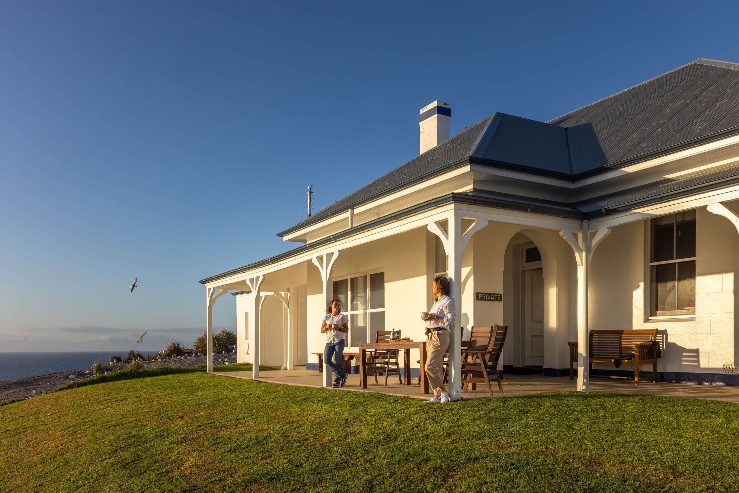 A house with a wraparound porch sits on a grassy hill overlooking the ocean. Two people stand on the porch, enjoying the view. The sky is clear and blue, and a few birds are flying nearby. A sign near the entrance reads "PRIVATE."