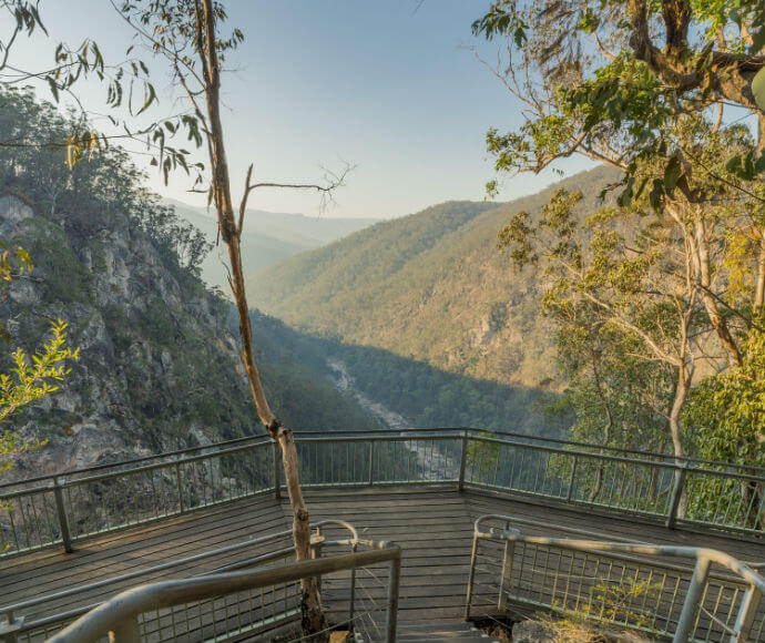 A scenic viewpoint with a wooden deck and metal railings overlooking a lush, green valley surrounded by forested hills. The deck has a tree growing through it and offers a panoramic view of the landscape, with a river or stream visible in the distance. The sky is clear, indicating a sunny day.