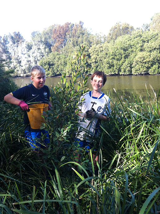 Two boys standing in grass with the Cooks River in the background holding pots from their work on bush regeneration.