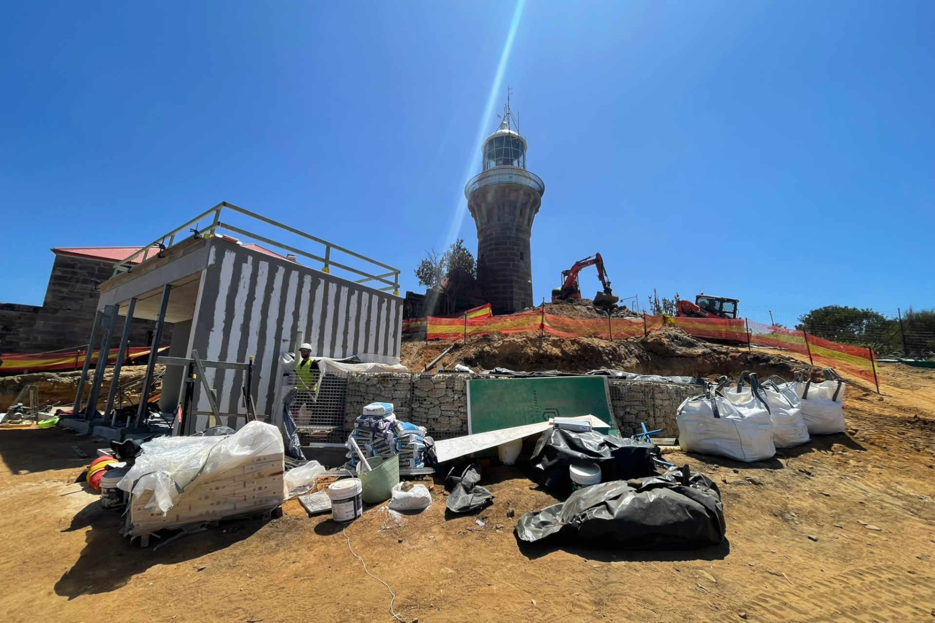 Construction materials and the shell of a new small building near the foot of a lighthouse under a wide blue sky