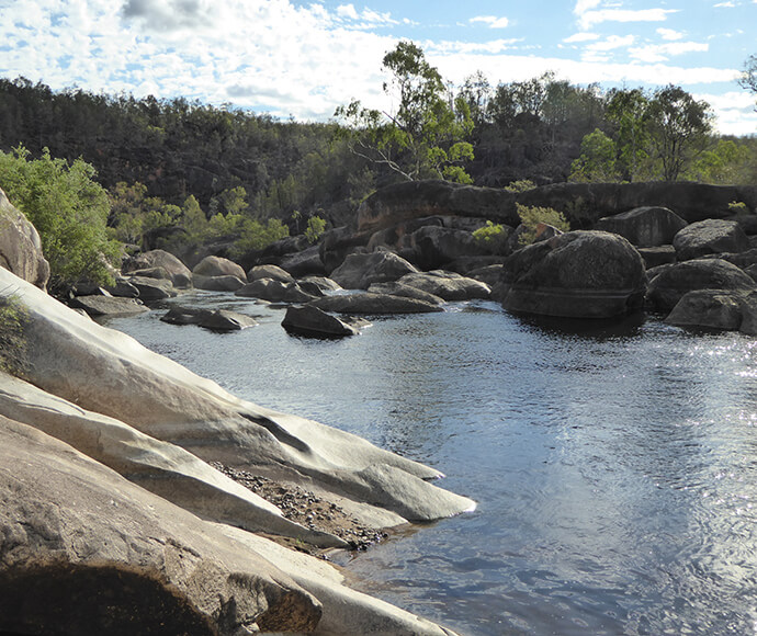 The image shows a natural landscape featuring a rocky river or stream. The water flows gently among large, smooth boulders and rocks. The surrounding area is lush with greenery, including trees and shrubs. The sky is partly cloudy, allowing sunlight to illuminate parts of the scene, creating reflections on the water's surface. The terrain appears rugged, with a mix of rocky outcrops and vegetation, suggesting a remote or wilderness area.