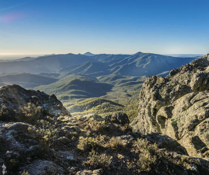 The image shows a scenic view of a mountainous landscape. The foreground features rocky terrain with some vegetation, while the background consists of multiple mountain ranges extending into the distance. The sky is clear and blue, indicating a sunny day.