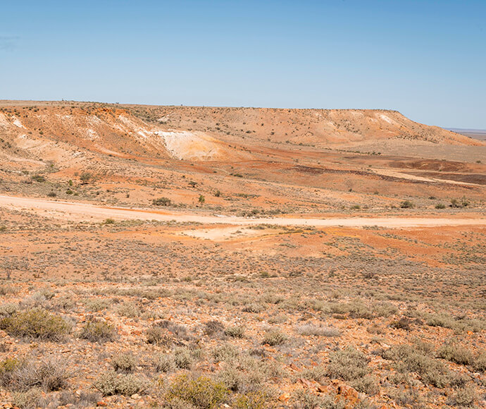 "A vast, arid landscape with sparse vegetation and a dirt road winding through it. The terrain is mostly flat with some low hills in the background under a clear blue sky.