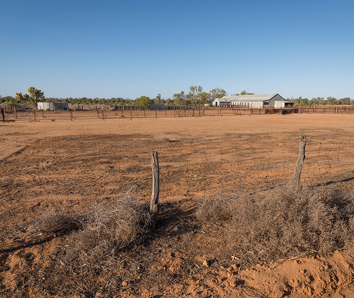 An image of the Outdoor Pastoral Museum in Corner Country, Sturt National Park, displaying how early European settlers worked on the land. The ground is mainly soil and stones with dried vegetation, and the area is fenced with wires and wooden poles. A large shed with a metal roof is visible at the back of the photo, all captured on a bright, clear sunny day.