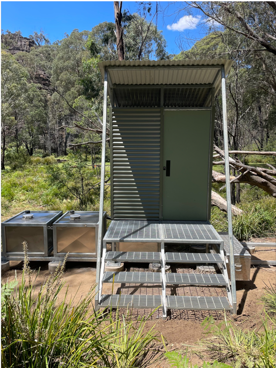 The toilet block is raised a few steps off the ground, and features two silver tanks to the left.