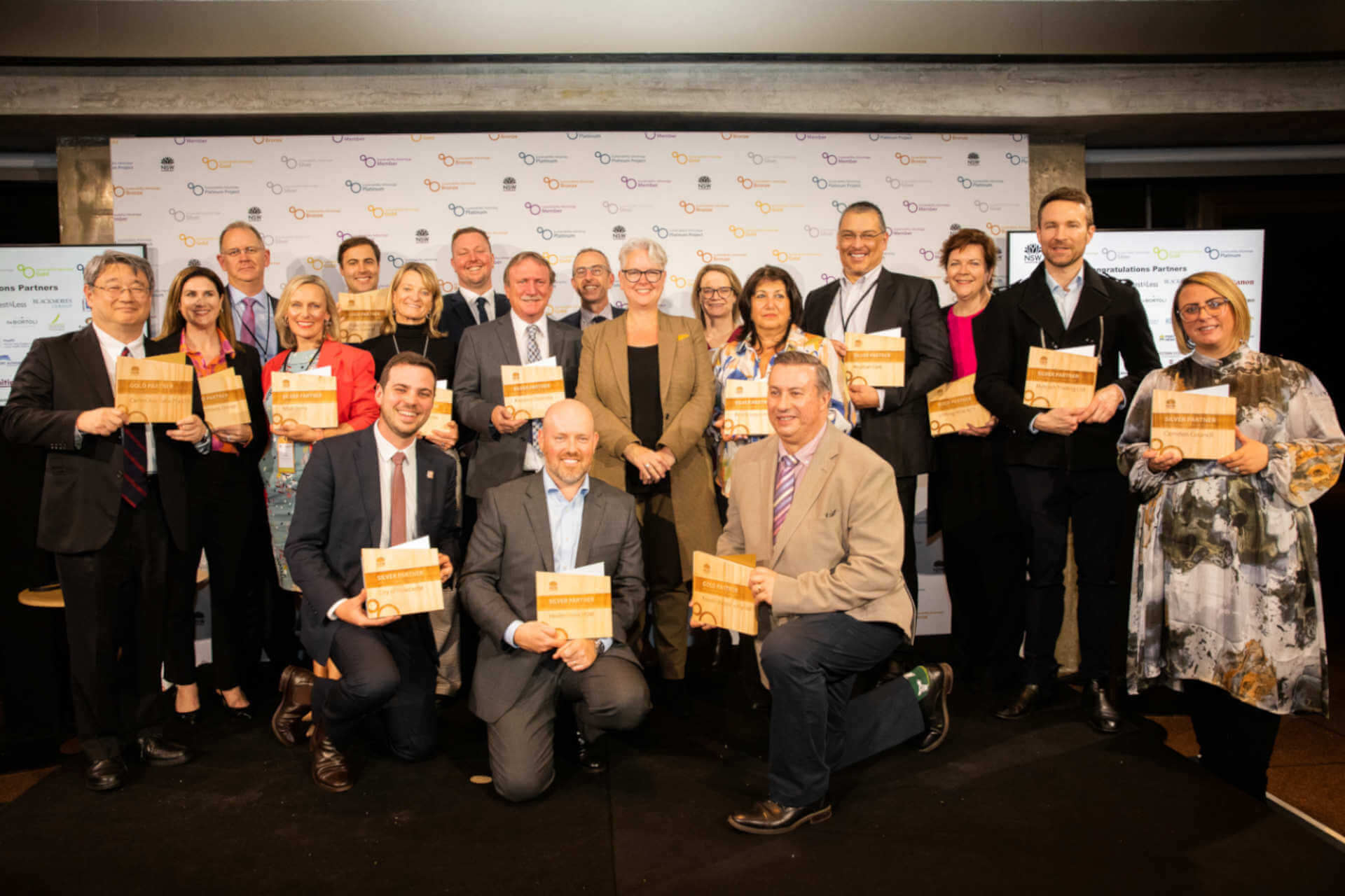 A group of smiling people semi-formally attired and holding awards in front of a NSW Government-branded screen