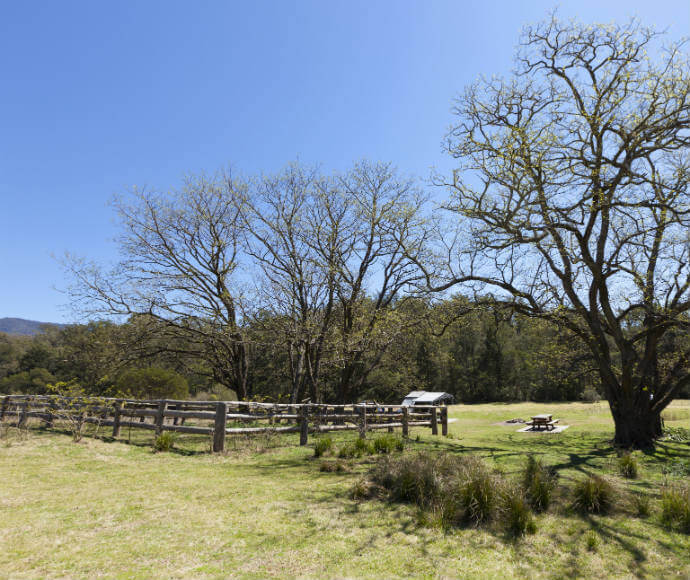 Photo of Bendethera Valley, Deua National Park. The image features a bright blue sky, a green grass lawn, wooden fences, and trees.