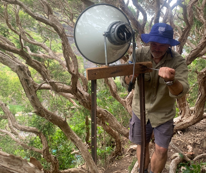 A man is using a screwdriver to mount a large speaker on a post in the bush