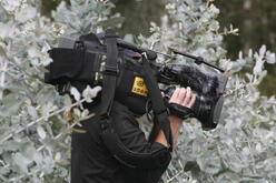 Man holding a camera on his shoulder standing next to a large shrub. 