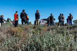 A group of people standing in a field with low shrubs under a clear blue sky, facing away from the camera towards a vehicle.