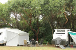 Camping site with two caravans and an annex surrounded by green trees, with empty chairs arranged outside, suggesting a peaceful outdoor vacation spot.