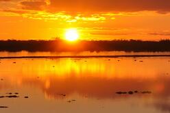 A vibrant orange sunset reflected in a calm body of water, with silhouetted land or vegetation lining the horizon.