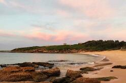 A curve of sand with a soft pink dawn, beach rocks in foreground