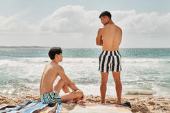 Two men in swimmersone sitting on a towel, the other is standing on sand, on the beach