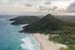 View of the coastline from above with ocean, a short stretch of sandy beach, hills and glimpses of sun through a cloudy sky in the distance.