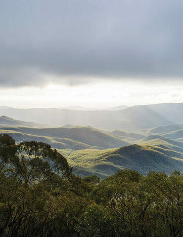 The image shows a scenic view of a mountainous landscape. The foreground features trees with dense foliage, while the background reveals a series of rolling hills and mountains extending into the distance. The sky is partly cloudy, with sunlight breaking through the clouds, casting rays of light onto the hills below. This interplay of light and shadow highlights the contours of the landscape, creating a visually striking and serene scene.