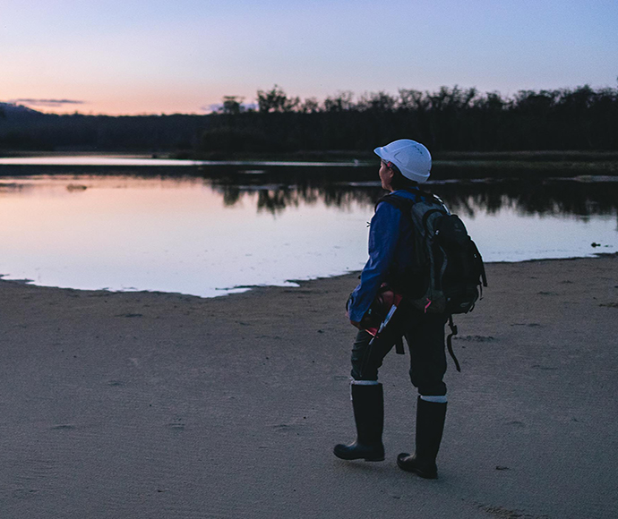 A researcher stands near water wearing gumboots and a hardhat and carrying a notebook