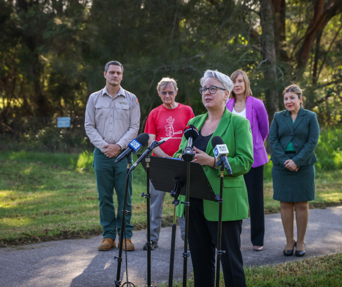 Penny Clarke, addressing the media behind a lectern and microphones outdoors in Wolli Creek Regional Park. Minister Sharpe is wearing a lime-green jacket. Behind her are 2 local politicians, an NPWS and a community group representative